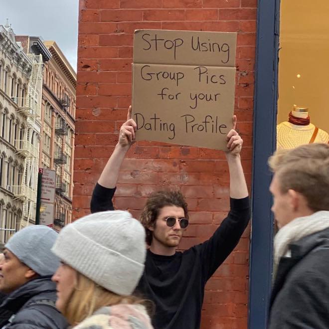 This meme features a photo of a person holding a protest-style sign in a crowded area. The sign reads "Stop using group pics for your dating profiles" and is likely being held by someone who is frustrated with others using group photos as their primary profile picture on dating apps. The image is meant to be humorous and relatable, poking fun at the common practice of using group photos on dating profiles.